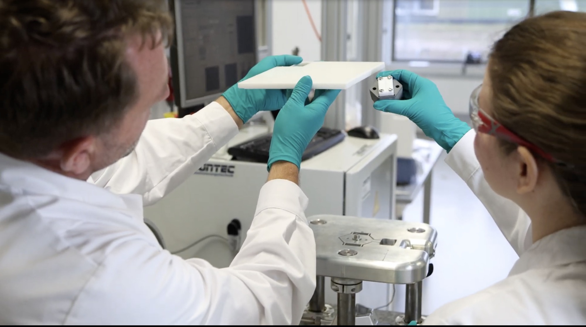 Two laboratory researchers examining a white panel sample beside testing equipment