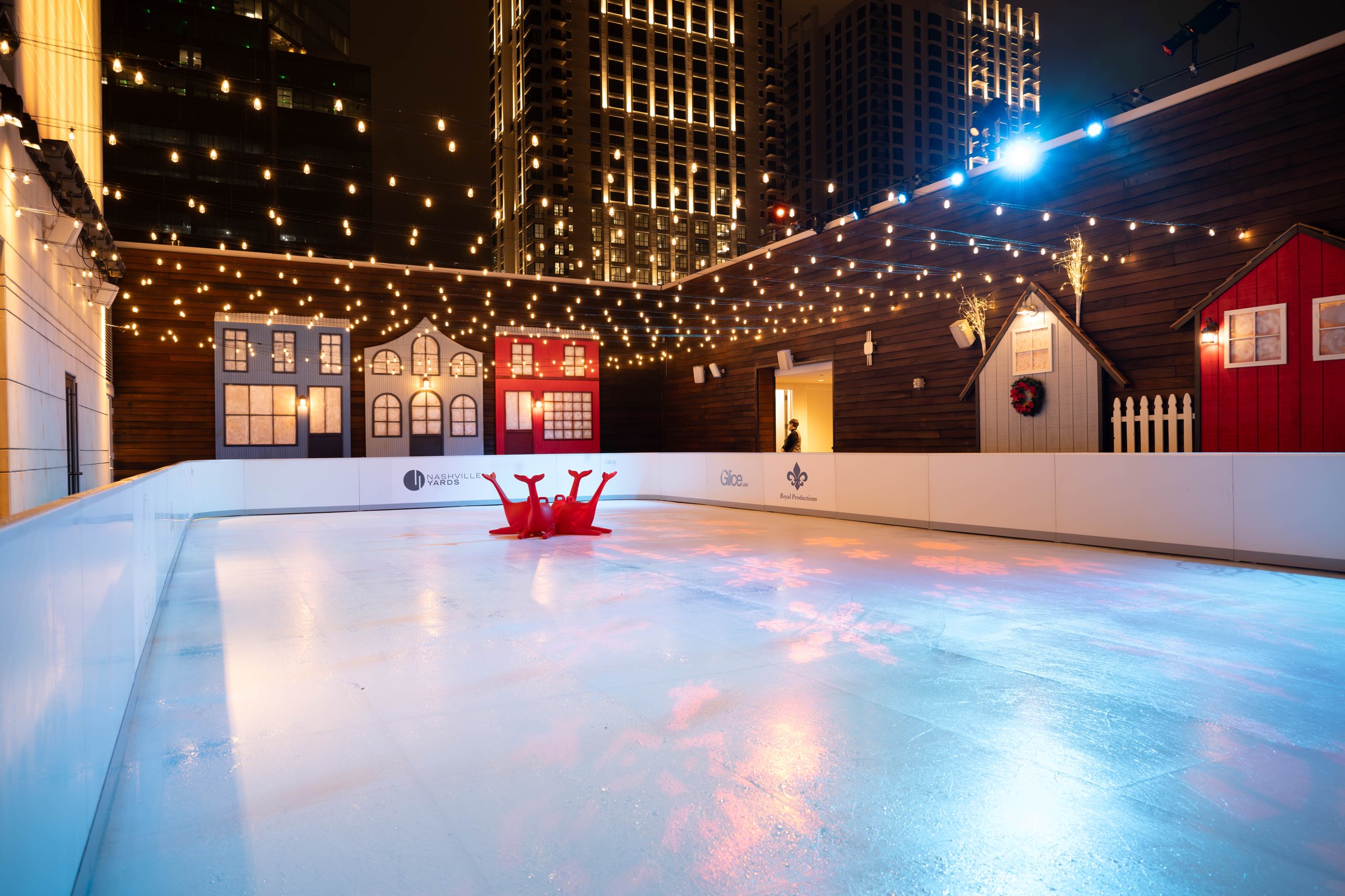 Empty Glice rink beneath string lights, with illuminated buildings in the background