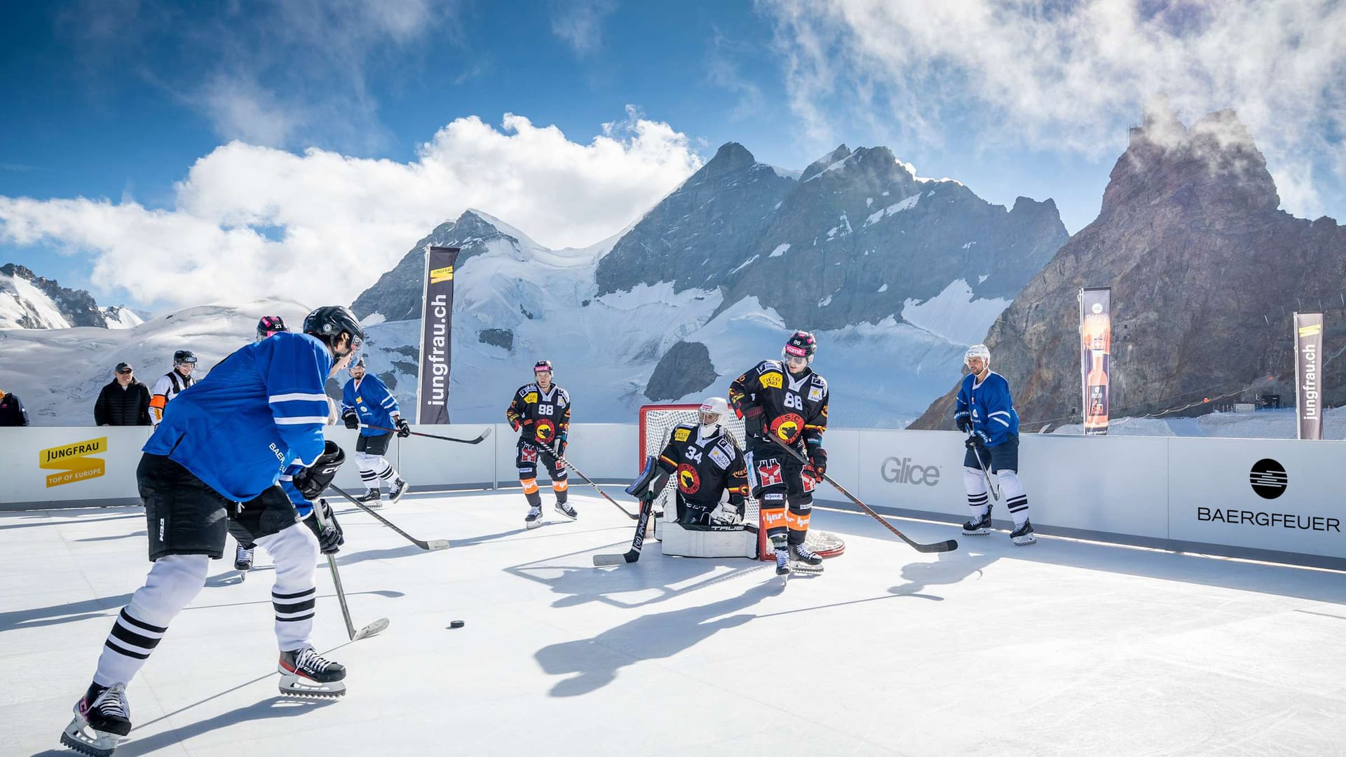 A hockey game on a compact boarded Glice rink outdoors at Jungfraujoch, players in front of the goal with snow-covered peaks behind the boards